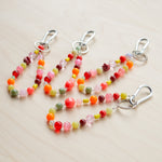 Three beaded keychains with silver metal clamps, featuring colorful round beads in pink, orange, red, yellow, green, and purple on a light wood background.