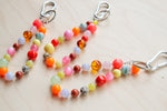 Two beaded keychain straps with silver carabiners, featuring multicolor round and faceted beads in pink, orange, yellow, green, brown, and white, laid out on a light wood surface.
